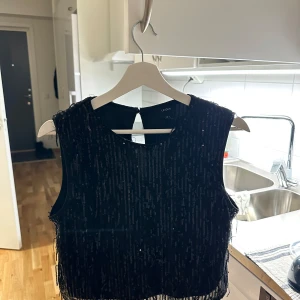 Svart glittrig ärmlös topp från Vero Moda - Snygg svart ärmlös topp från Vero Moda med glittriga detaljer. Perfekt för en kväll ute eller festliga tillfällen. Toppen har en rund halsringning och en elegant design.