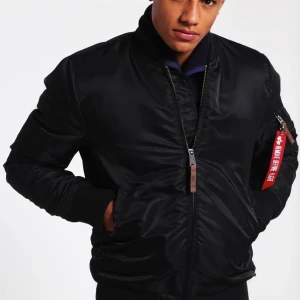 Svart Alpa industries jacka  -  Svart bomberjacka från Alpha Industries med klassisk dragkedja och 'Remove Before Flight'-tag på ärmen. Jackan har en orange insida och ribbade muddar vid ärmslut och midja. Använd fåtal gånger 