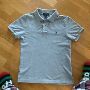 Grå pikétröja från Ralph Lauren - Säljer en klassisk grå pikétröja från Ralph Lauren. Tröjan är grå med ett svart märke. Den är storlek L i barn och passar dig som är runt 170 perfekt!