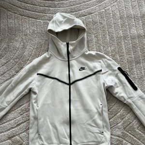  Nike tech Cream white kofta - Snygg vit hoodie från Nike tech med dragkedja och huva. Svarta detaljer och logga på bröstet. Praktisk ficka med dragkedja på ärmen. Perfekt för en sportig look.