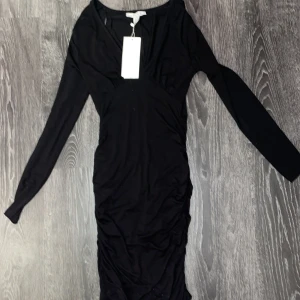 Svart klänning med v-ringning - Elegant svart klänning med v-ringning och långa ärmar. Perfekt för en stilren look. Klänningen har en figurnära passform som framhäver silhuetten. Den sitter tajt på kroppen med en urringning upp till.