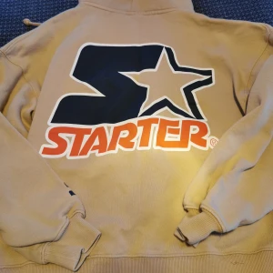 Beige hoodie från Starter/ Pull&Bear - Säljer en beige hoodie från Starter med stor logga på ryggen och mindre logga på bröstet. Tröjan har dragkedja och huva, samt ribbade muddar vid ärmslut. Perfekt för en avslappnad stil.