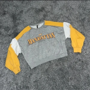Grå och gul sweatshirt - Snygg grå sweatshirt med texten 'Manhattan' på bröstet. Tröjan har långa ärmar i gult och vitt. Perfekt för en avslappnad stil.