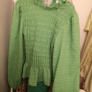 Grön blus med volangdetaljer - Snygg grön blus med volangdetaljer och smockad design. Blusen har långa ärmar och en luftig passform, perfekt för vår och sommar. Den unika strukturen ger en trendig look.