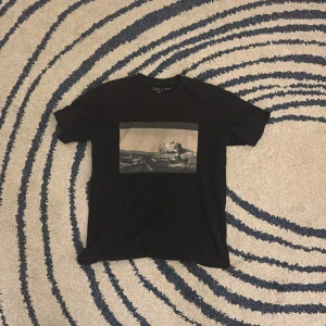 Svart t-shirt med tryck från Uniqlo - Snygg svart t-shirt från Uniqlo med ett coolt tryck på framsidan och texten 'United States, Wyoming 1974' på baksidan. Perfekt för en avslappnad stil. Lite slitage på etiketten men inget synligt slitage allt när den används. Foto på trycket av Elliot erwitt