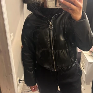 Svart pufferjacka med dragkedja - Snygg svart pufferjacka med dragkedja framtill. Jackan har en hög krage och långa ärmar, perfekt för kyligare dagar. Den har en glansig yta som ger en modern look.