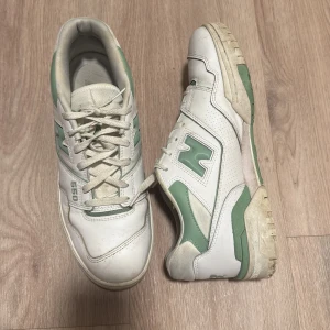 Vita och gröna sneakers från New Balance - Säljer ett par vita sneakers från New Balance med gröna detaljer och snörning. Modellen är 550 och har en klassisk design med bekväm passform. Perfekta för en avslappnad stil.