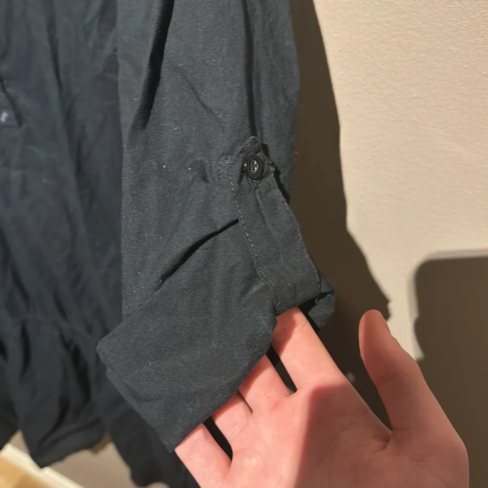 Snygg svart klänning från Gina Tricot i bomull. Den har trekvartsärmar med knappdetalj och en v-ringad hals. Perfekt för en stilren look.. Mekot.