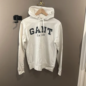 Vit hoodie från GANT - Säljer en vit hoodie från GANT med tryck på framsidan. Den har en klassisk design med huva och snören för att kunna dra åt huvan. Perfekt för en avslappnad stil. Det ska dock tilläggas att det finns en större fläck på baksidan då plagget är använt ett stort antal gånger. Storleken är M men sitter som S