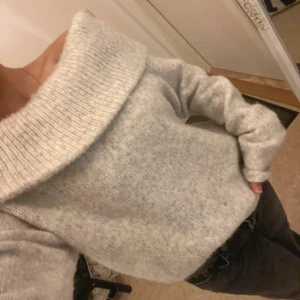 Grå offshoulder stickad tröja - Säljer en mysig grå stickad tröja med offshoulder-design. Tröjan har långa ärmar och en bred ribbad kant vid axlarna som ger en stilren look. Perfekt för kyligare dagar!