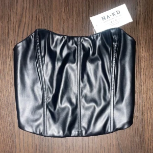 Svart korsett från NA-KD - Snygg svart korsett i skinnimitation från NA-KD. Perfekt för en edgy look med en tight passform. Korsetten har en stilren design utan axelband och är lätt att matcha med olika outfits. LÄGGER UPP FÖR FRIFRAKT