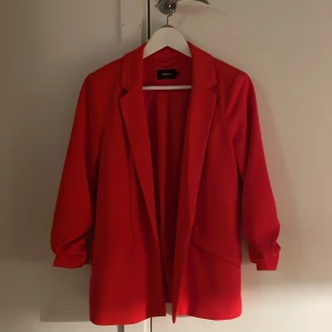 Röd kavaj från ONLY - Snygg röd kavaj från ONLY med detalj i armarna och stilren design. Perfekt för att liva upp garderoben med en färgklick. Kavajen har en klassisk krage och är tillverkad i ett bekvämt material.