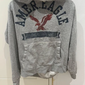 Grå hoodie från American Eagle - Säljer en grå hoodie från American Eagle med tryck av en örn och texten 'AMER. EAGLE' på framsidan. Tröjan har en stor ficka framtill och en bekväm huva. Perfekt för en avslappnad stil.