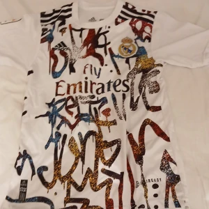 Vit fotbollströja med grafiskt mönster - Snygg vit fotbollströja från Adidas med ett unikt grafiskt mönster i flera färger. Tröjan har korta ärmar och Real Madrids klubbmärke på bröstet. Perfekt för fotbollsfans som vill sticka ut!