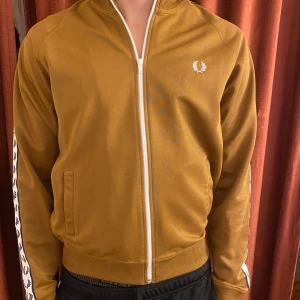 Brun track jacket från Fred Perry - Säljer en snygg brun track jacket från Fred Perry med vita detaljer. Jackan har en dragkedja framtill och ribbade muddar. Ärmarna pryds av ett vitt band med Fred Perry-loggan. Modell 185 cm 