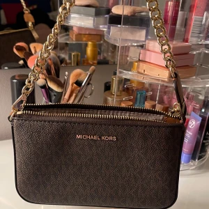 Brun axelväska från Michael Kors - Snygg brun axelväska från Michael Kors med guldfärgad kedja och dragkedja. Perfekt för att bära dina nödvändigheter med stil. Väskan har en elegant design och är tillverkad i syntetmaterial. Använd fåtal gånger och köpt för 1800kr🤎