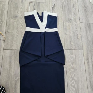 Marinblå klänning med vita detaljer - Elegant marinblå klänning med vita kontrastdetaljer och v-ringning. Klänningen har smala axelband och en figurnära passform som framhäver silhuetten. Perfekt för en stilfull kväll.