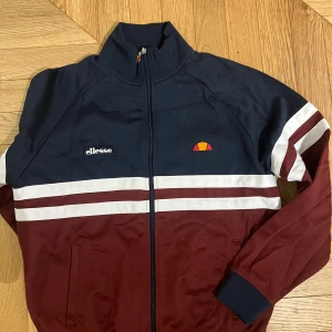 Blå och vinröd jacka från Ellesse - Snygg blå och vinröd jacka från Ellesse med dragkedja och vita ränder. Jackan har långa ärmar och en klassisk sportig look med broderad logotyp på bröstet.