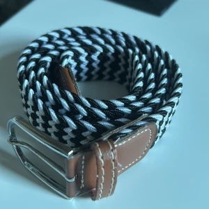 Svart och vitt flätat bälte med brunt läder - Snyggt flätat bälte i svart och vitt med detaljer i brunt läder. Perfekt för att ge en stilren touch till din outfit. Spännet är i silverfärgad metall. Kommer ej till användning längre och vill ha något nytt bäste istället. 