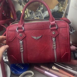 Zadig handväska - !!ej äkta!! Snygg röd handväska i skinn med två dekorativa dragkedjor framtill och ett litet emblem. Väskan har både handtag och en avtagbar axelrem för flexibilitet. Perfekt för att ge en färgklick till din outfit.