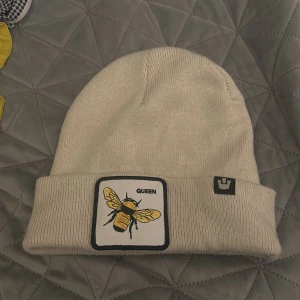 Vit stickad mössa med bi-märke - Snygg vit stickad mössa med ett broderat bi-märke och texten 'QUEEN' framtill. Perfekt för att hålla värmen med stil.
