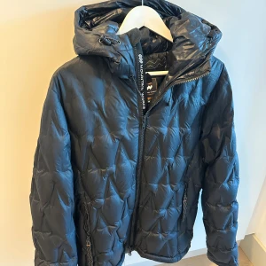 Svart dunjacka från Mountain Works  - Snygg svart dunjacka från  Mountain Works med quiltad design och praktisk huva. Jackan har dragkedja framtill och är perfekt för kyliga dagar. Den är både stilren och funktionell. Jackan är sparsamt använd. 