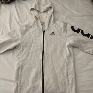 Vit huvtröja från Adidas - Säljer en vit huvtröja från Adidas med dragkedja och svart logga på ärmen. Perfekt för en sportig look eller avslappnade dagar. Tröjan har långa ärmar och en bekväm huva.