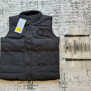 Svart dunväst från Canada Goose - Säljer en stilren svart dunväst från Canada Goose med knappar och två stora fickor framtill. Västen har en rund logotyp på bröstet och är perfekt för kyligare dagar. Den är fodrad och har en hög krage för extra värme.