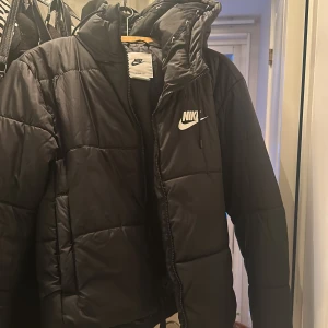 Svart dunjacka från Nike - Säljer en svart dunjacka från Nike med Therma-Fit teknologi. Jackan har en dragkedja framtill och en huva med små Nike-loggor längs kanten. Perfekt för kyliga dagar.