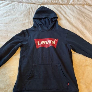 Marinblå hoodie från Levi's - Säljer en marinblå hoodie från Levi's med det klassiska röda logotyptrycket på framsidan. Tröjan har en stor ficka framtill och en bekväm huva. Perfekt för en avslappnad stil.