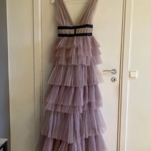 Lila volangklänning med djup urringning - Använd en gångSäljer en fantastisk lila långklänning med flera lager av volanger och en djup v-ringad urringning. Klänningen har svarta band i midjan som ger en snygg kontrast och markerar figuren. Perfekt för speciella tillfällen!
