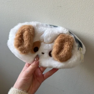 Vit pälsig plånbok med hundmotiv - Söt och fluffig plånbok i vitt med ett gulligt hundansikte och bruna öron. Den har en praktisk dragkedja för säker förvaring av dina saker. Perfekt för att lägga till en lekfull touch till din vardag.
