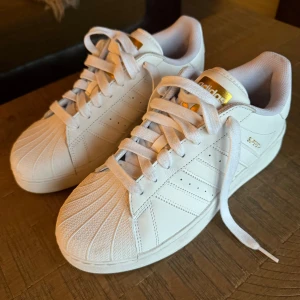 Vita Adidas Superstar sneakers - Snygga vita Adidas Superstar sneakers med klassisk snäckformad tå och gyllene detaljer. Skorna har snörning och den ikoniska tre-randiga designen på sidorna. Perfekta för en stilren look. Skorna är nya och bara använda 1 gång. Vid frågor är det bara att skriva MVH/Felix