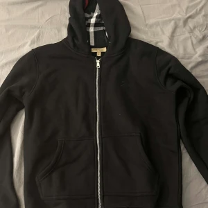 Svart huvtröja från Burberry - Snygg svart huvtröja från Burberry med dragkedja och klassiskt rutigt mönster på insidan av huvan. Perfekt för en stilren look med en touch av lyx. Tröjan har långa ärmar och praktiska fickor framtill.