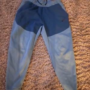 Nike tech byxor - Snygga blå mjukisbyxor från Nike med mörkare partier upptill. Byxorna har en bekväm passform med snörning i midjan och är perfekta för en avslappnad stil. Perfekta för både träning och chill.