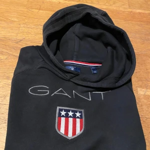 Svart hoodie från GANT - Snygg svart hoodie från GANT med broderad logga och emblem på bröstet. Tröjan har en klassisk känguruficka och en bekväm huva. Perfekt för en avslappnad stil. Stl 170 