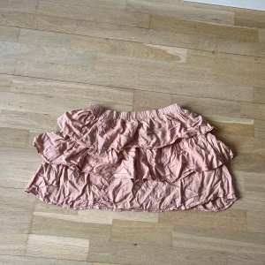 Rosa volangkjol - Söt rosa kjol med flera lager volanger som ger en lekfull look. Perfekt för varma dagar med sin korta längd och lätta material. Den elastiska midjan gör den bekväm att bära hela dagen.