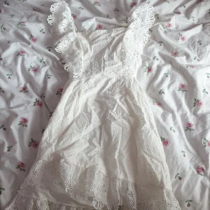 Vit spetsklänning med volangdetaljer - Charmig vit klänning med vackra spetsdetaljer och volanger. Klänningen har en öppen rygg och breda axelband med spets. Perfekt för sommarens festligheter.