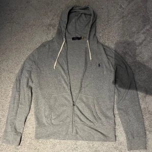 Grå hoodie från Ralph Lauren - Säljer en stilren grå zip hoodie från Ralph Lauren med dragkedja. Perfekt för en avslappnad look. Tröjan är äkta 