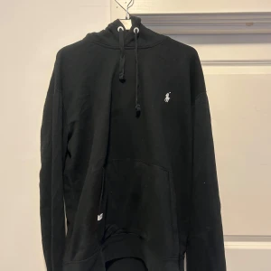 Svart hoodie från Ralph Lauren Large - Säljer en klassisk svart hoodie från Ralph Lauren med det ikoniska logotypbroderiet på bröstet. Tröjan har en justerbar huva med snören och en stor magficka framtill. Perfekt för en avslappnad stil.