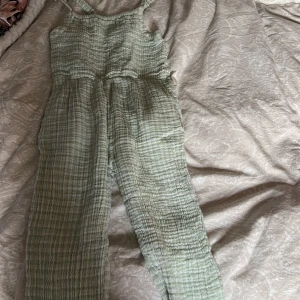 Grön randig jumpsuit från Lindex - Säljer en söt grön randig jumpsuit från Lindex. Den har smala axelband och en smockad överdel som ger en bekväm passform. Perfekt för varma dagar med sitt lätta och luftiga material. 🌿