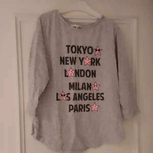 Grå långärmad tröja till flicka med städer, storlek 12 år,152 - Snygg grå långärmad tröja från H&M med texten Tokyo, New York, London, Milan, Los Angeles och Paris i svart. Varje stad är dekorerad med en rosa stjärna. Perfekt för en avslappnad stil. Det är storlek 12 år,152