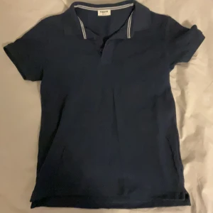 Mörkblå pikétröja från Kappahl - Säljer en stilren mörkblå pikétröja från Kappahl. Tröjan har korta ärmar och en klassisk krage med vita ränder. Perfekt för en avslappnad look.