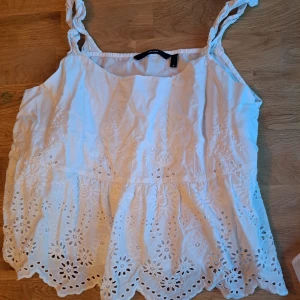 Vit broderad linne från H&M - Charmig vit linne från H&M med broderade blommönster och spetsdetaljer. Den har knytbara axelband och en luftig passform, perfekt för varma sommardagar.