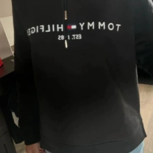 Svart hoodie från Tommy Hilfiger - Säljer en stilren svart hoodie från Tommy Hilfiger med dragkedja och logga framtill. Tröjan har en klassisk design med huva och dragsko. Perfekt för en avslappnad look.