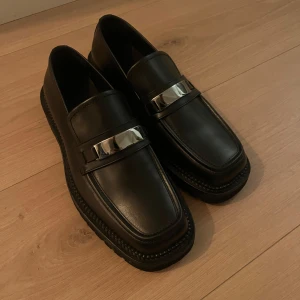 Svarta loafers med metallspänne - Stilrena svarta loafers från Filippa K (nypris ca 4900) med ett elegant metallspänne på ovansidan. Skorna har en robust sula och är tillverkade i skinn, vilket ger en klassisk look. Perfekta för en stilren och modern outfit. Storleken är 42 men passar mer som 43!
