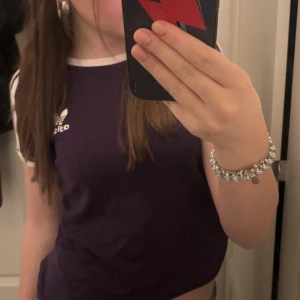 Lila t-shirt från Adidas - Säljer en snygg lila t-shirt från Adidas med vita detaljer och klassiska ränder på ärmarna. T-shirten har en rund halsringning och korta ärmar. Perfekt för en sportig look!