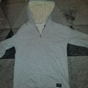 Grå hoodie med teddyfoder - Mysig grå hoodie med teddyfoder i luvan. Perfekt för kyliga dagar. Har en dragkedja vid halsen och en praktisk magficka framtill.
