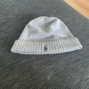 Grå mössa från Polo Ralph Lauren - Snygg grå mössa från Polo Ralph Lauren i 100% merinoull. Den har en ribbad kant och en broderad logga framtill. Perfekt för kyliga dagar.