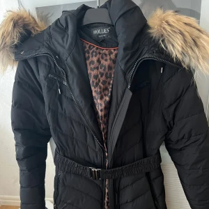 Vinterjacka Hollies - Snygg svart dunjacka från Hollies med pälskantad huva och bälte i midjan. Perfekt för kallare dagar. 